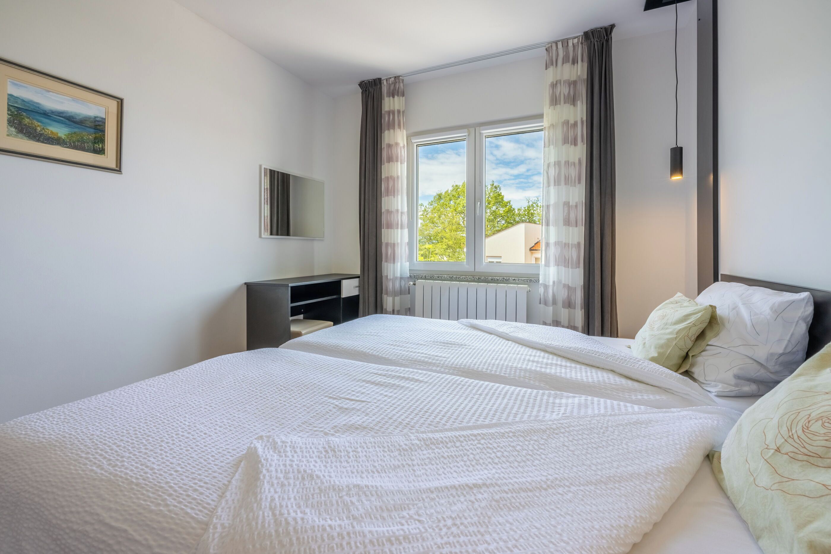 A bedroom with a large bed, a window with curtains, a bedside table, and a painting on the wall.
