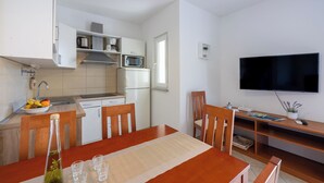 A compact kitchen with white cabinets, a microwave, and a refrigerator. There is a dining table with chairs, a TV mounted on the wall, and a small shelf with a plant.