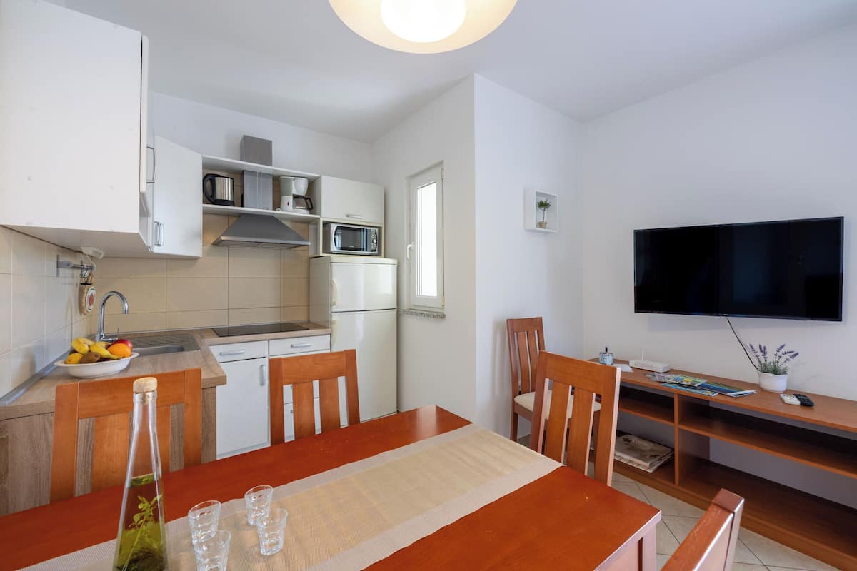 A compact kitchen with white cabinets, a microwave, and a refrigerator. There is a dining table with chairs and a TV mounted on the wall.
