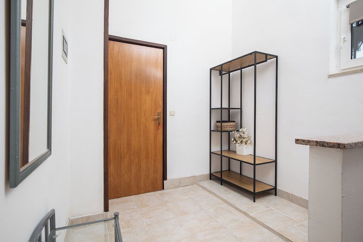 A hallway with a wooden door, a metal shelving unit with baskets and flowers, and a tiled floor.
