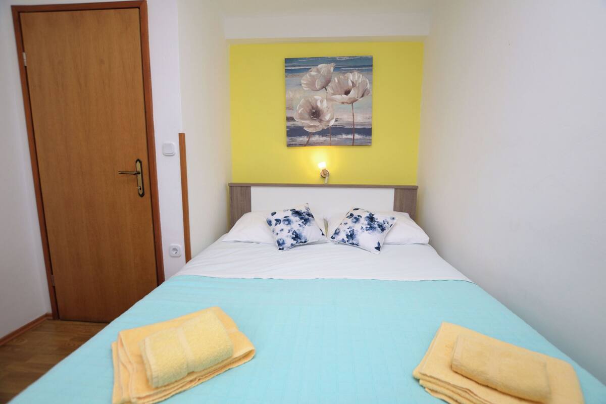 A hotel room with a bed, a yellow wall, and a painting of flowers.