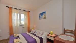 Un dormitorio con una cama, dos mesitas de noche, una silla de madera, una ventana con cortinas y un cuadro colgado en la pared.