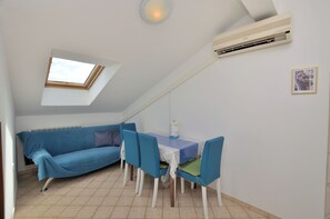 Et lille, hyggeligt rum med skråvægge, et ovenlysvindue, en blå sofa, et spisebord med stole og en aircondition.