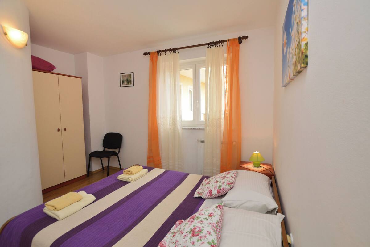 A bedroom with a bed, a chair, a wardrobe, a window with curtains, and a lamp.