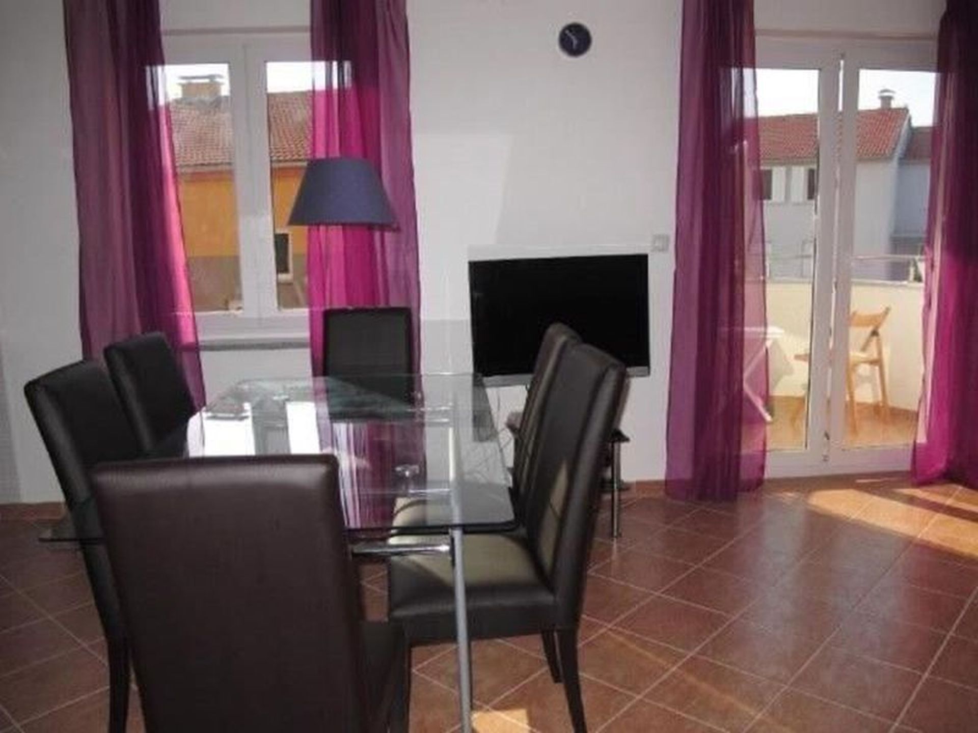 A dining area with a glass table, four chairs, a TV, and a sliding glass door leading to a balcony.