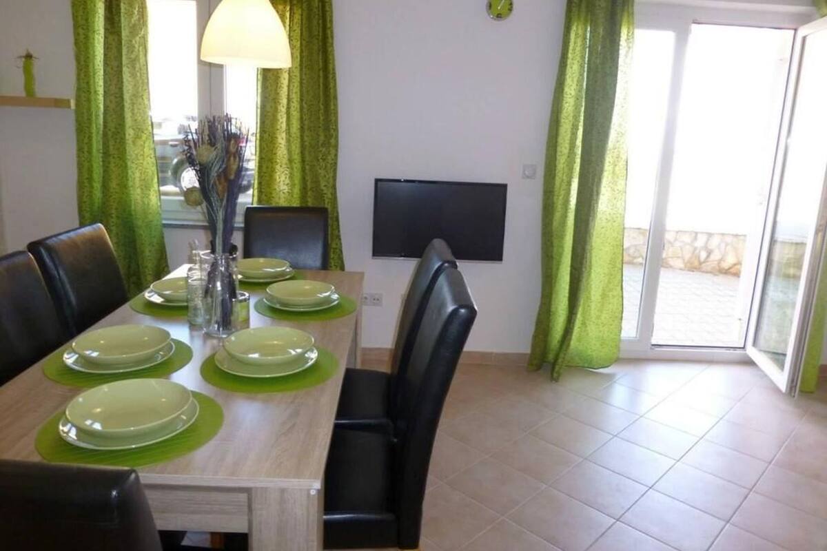 A dining area with a table set for four, green curtains, a TV on the wall, and a view through the glass door.