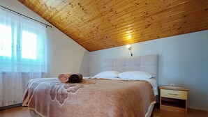 A bedroom with a wooden ceiling, a bed with a pink blanket, two rolled towels, white pillows, a nightstand, and a window with sheer curtains. - Apartments Marica (Cizici)