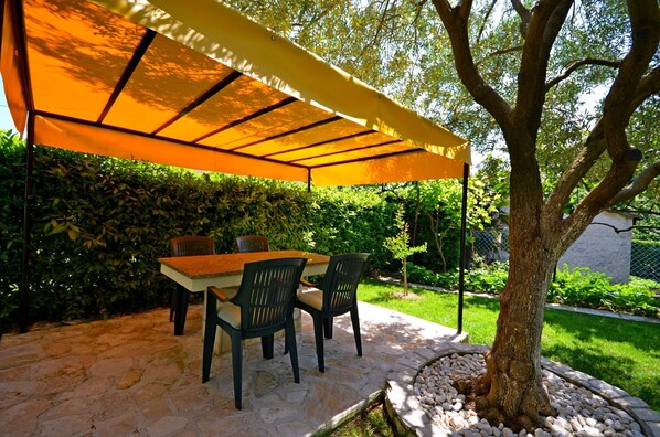 An outdoor patio with a table and chairs under a canopy, surrounded by greenery and a stone pathway.