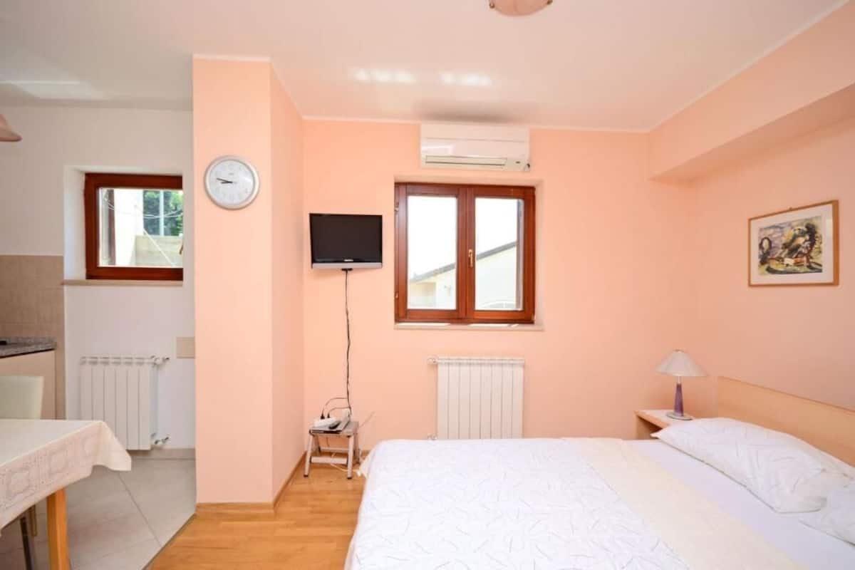 A bedroom with a bed, a TV, a window, a radiator, and a table with a chair.