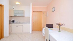 A compact kitchen with white cabinets, a microwave, and a sink. There's a dining table with a vase of flowers and a clock on the wall. - Apartments Ivanka (Senj)
