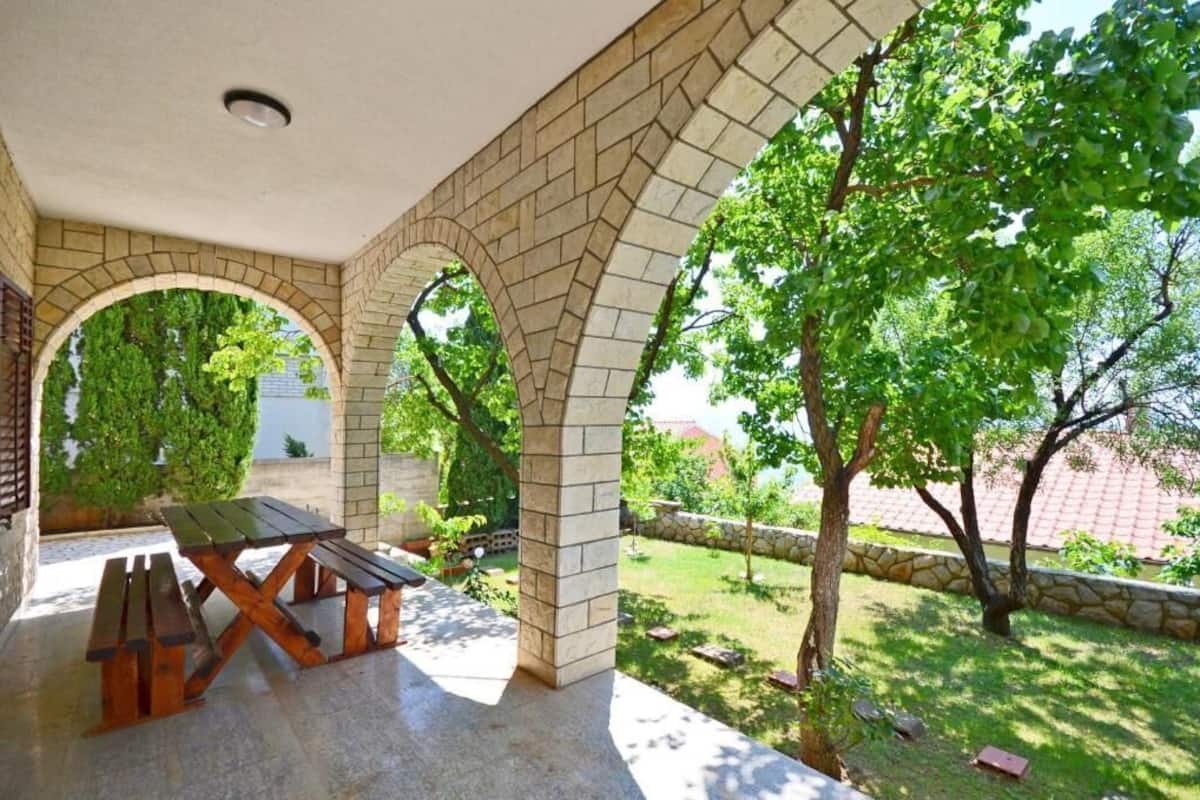 A covered patio with a wooden table and benches, surrounded by brick arches and greenery.