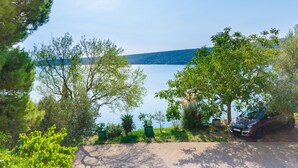 A lakeside view with a car parked under a tree, surrounded by greenery and a clear sky. - Apartments Ratko (Rakalj)