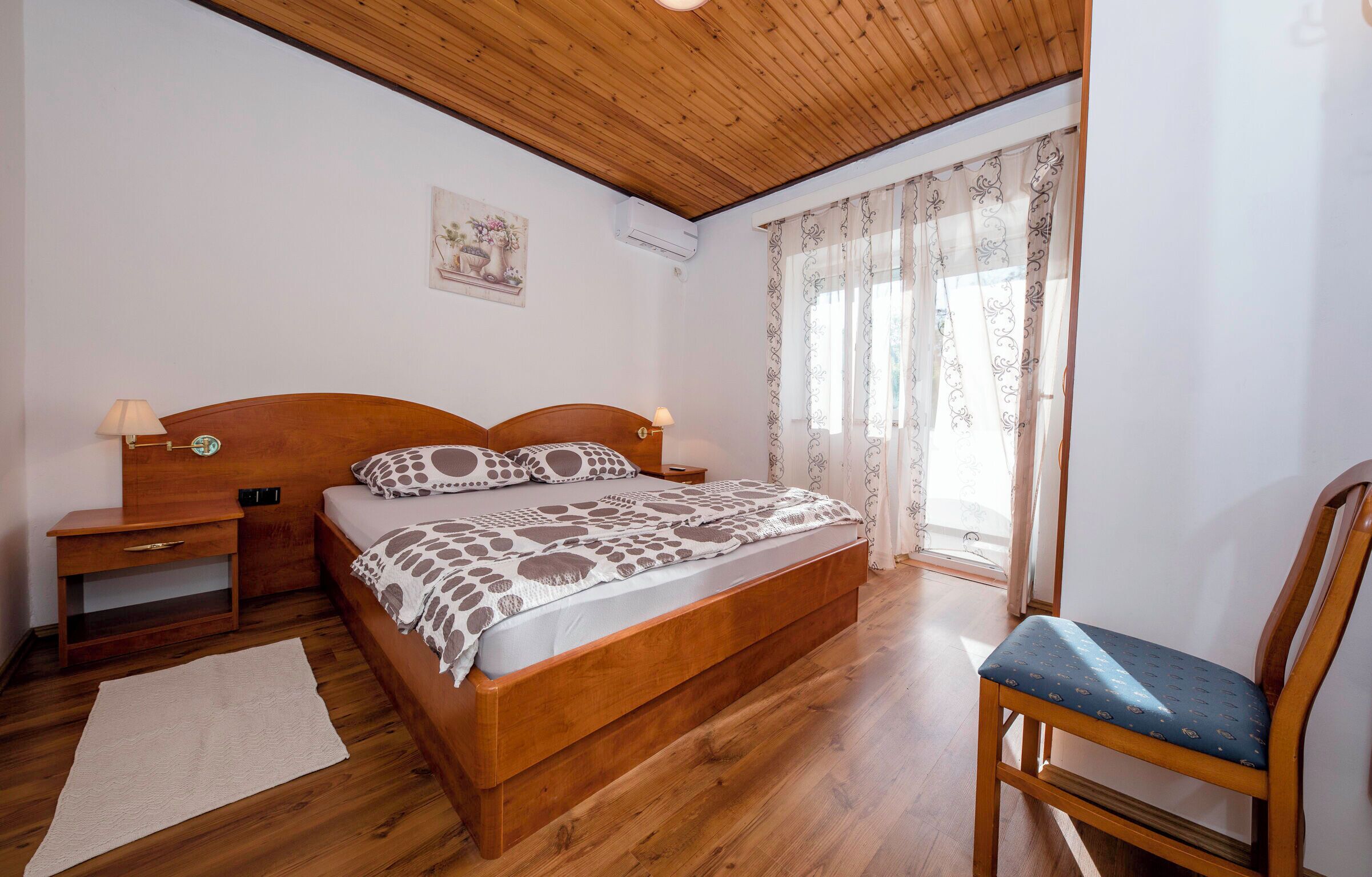 Une chambre à coucher avec un lit en bois, des tables de chevet, une chaise, une fenêtre avec des rideaux et un climatiseur fixé au mur.