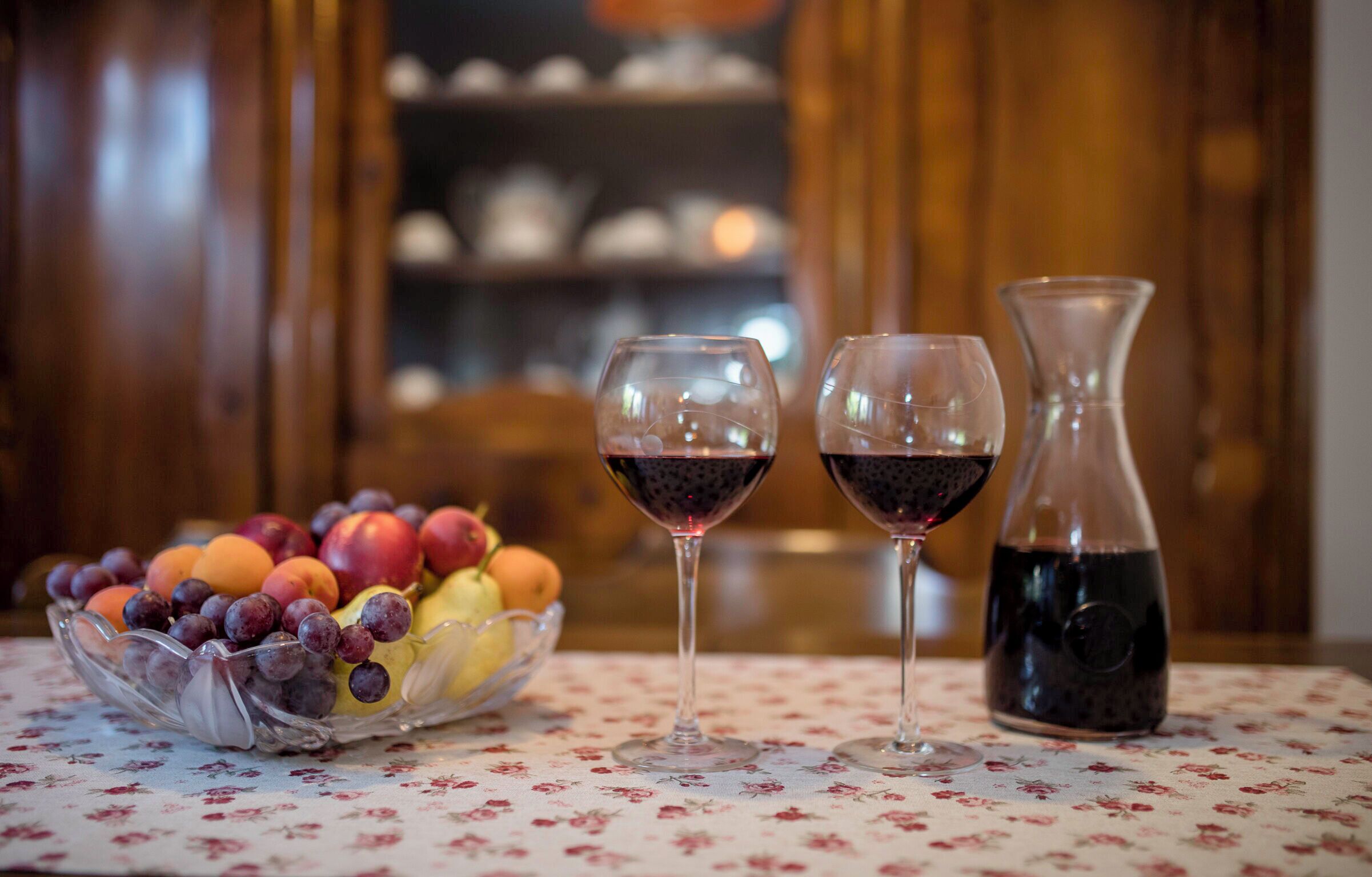 A table with a floral tablecloth, a bowl of fruit, and two wine glasses with a bottle of red wine.