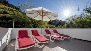 A sunlit outdoor seating area with white lounge chairs and red cushions, an umbrella, and a view of a hillside. - House Predrag (Supetarska Draga)