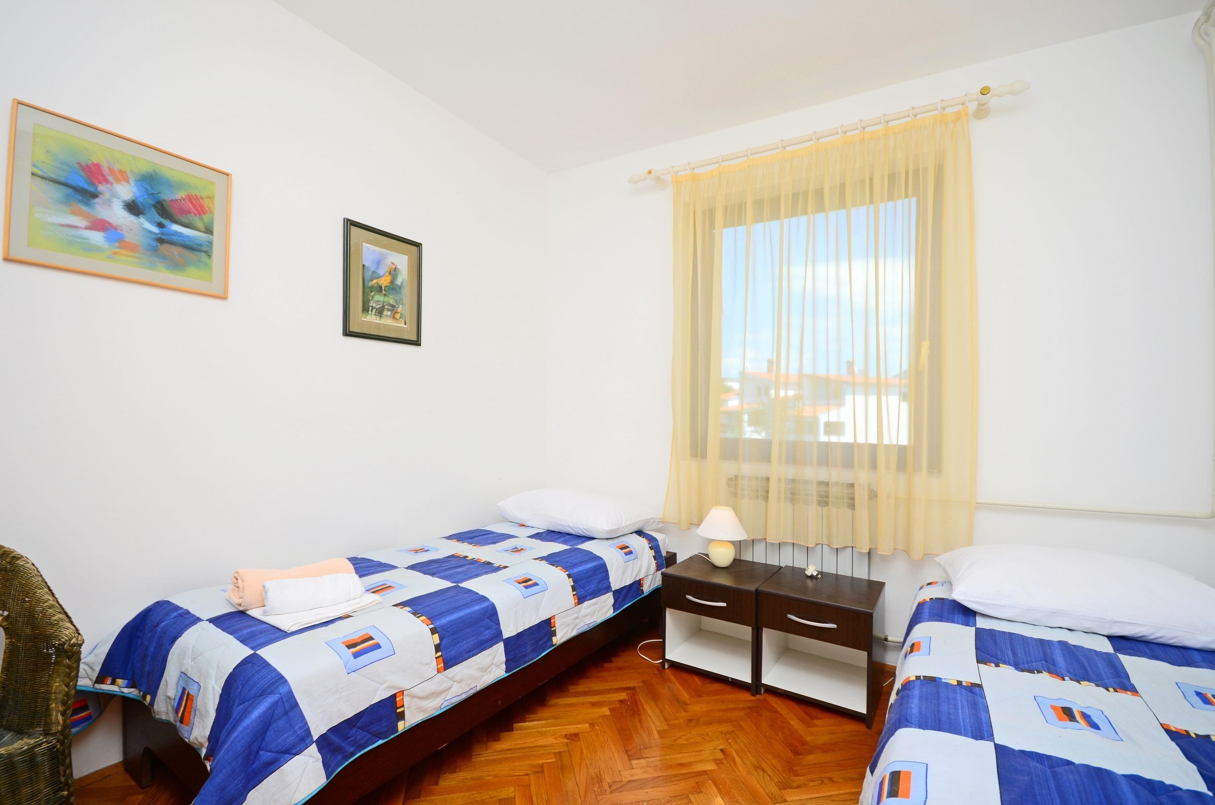 A room with two beds, a wooden floor, a small desk, a chair, and a window with curtains.