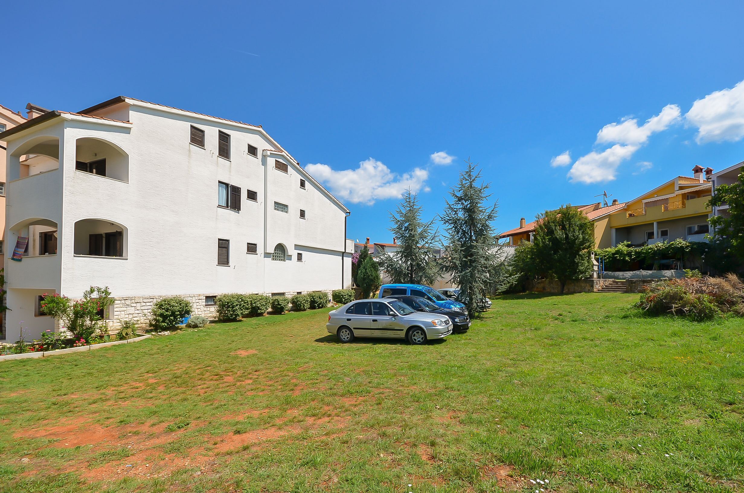 Un edificio residencial con fachada blanca, múltiples ventanas y techo plano. Hay dos coches estacionados en el jardín delantero, y la zona está rodeada de vegetación y árboles.