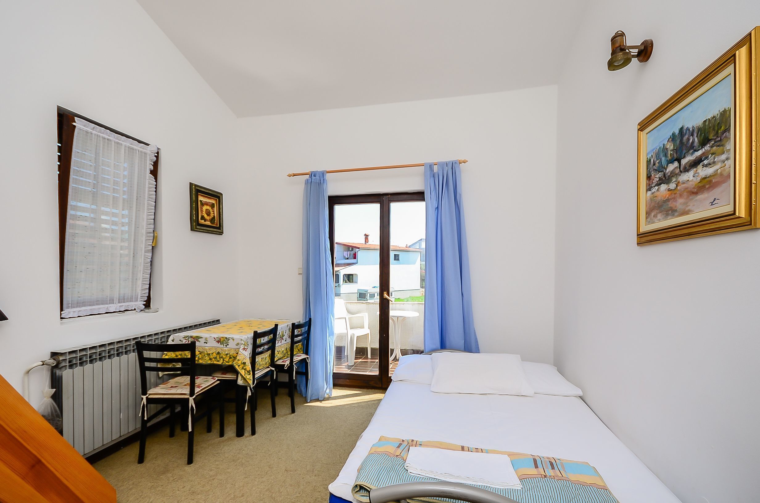 Habitación de hotel con cama, una mesa con sillas, un radiador, una ventana con cortinas y una puerta que da a un espacio exterior.