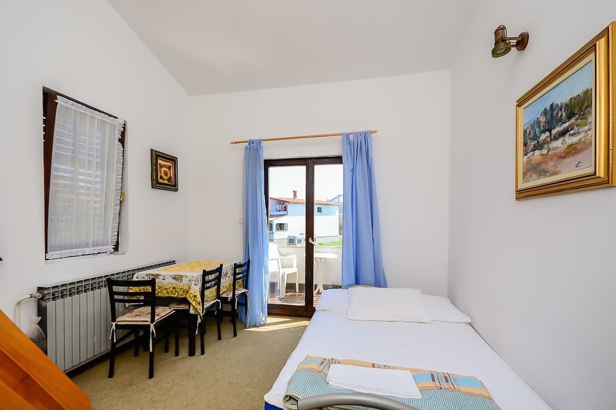 Habitación de hotel con cama, una mesa con sillas, un radiador, una ventana con cortinas y una puerta que da a un espacio exterior.