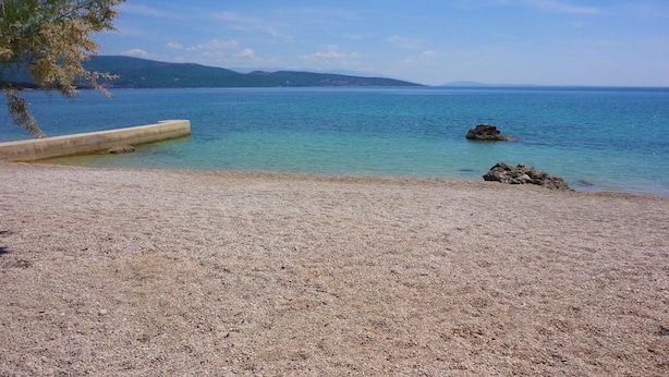 Ein Kiesstrand mit klarem Wasser, einer Betonbarriere und fernen Bergen.