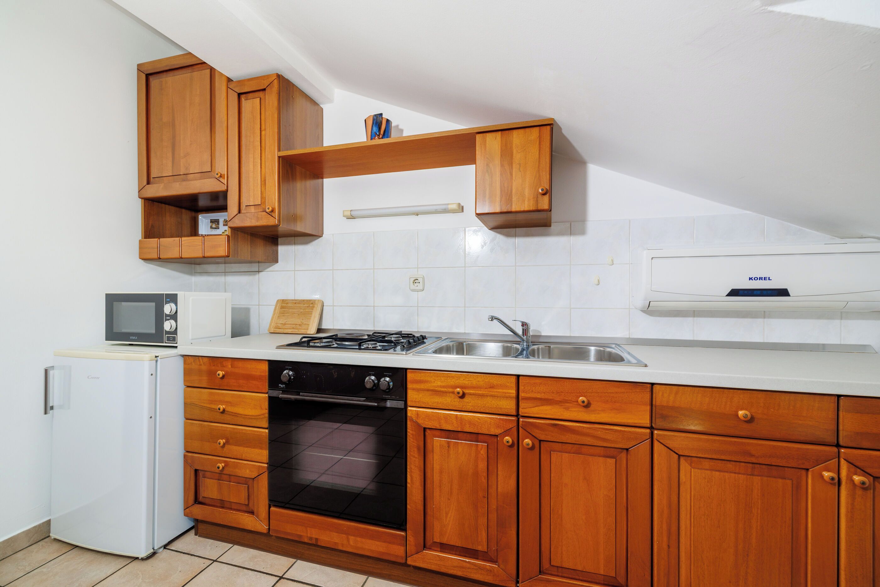 Een kleine keuken met houten kastjes, een magnetron, een kookplaat en een ingebouwde oven.