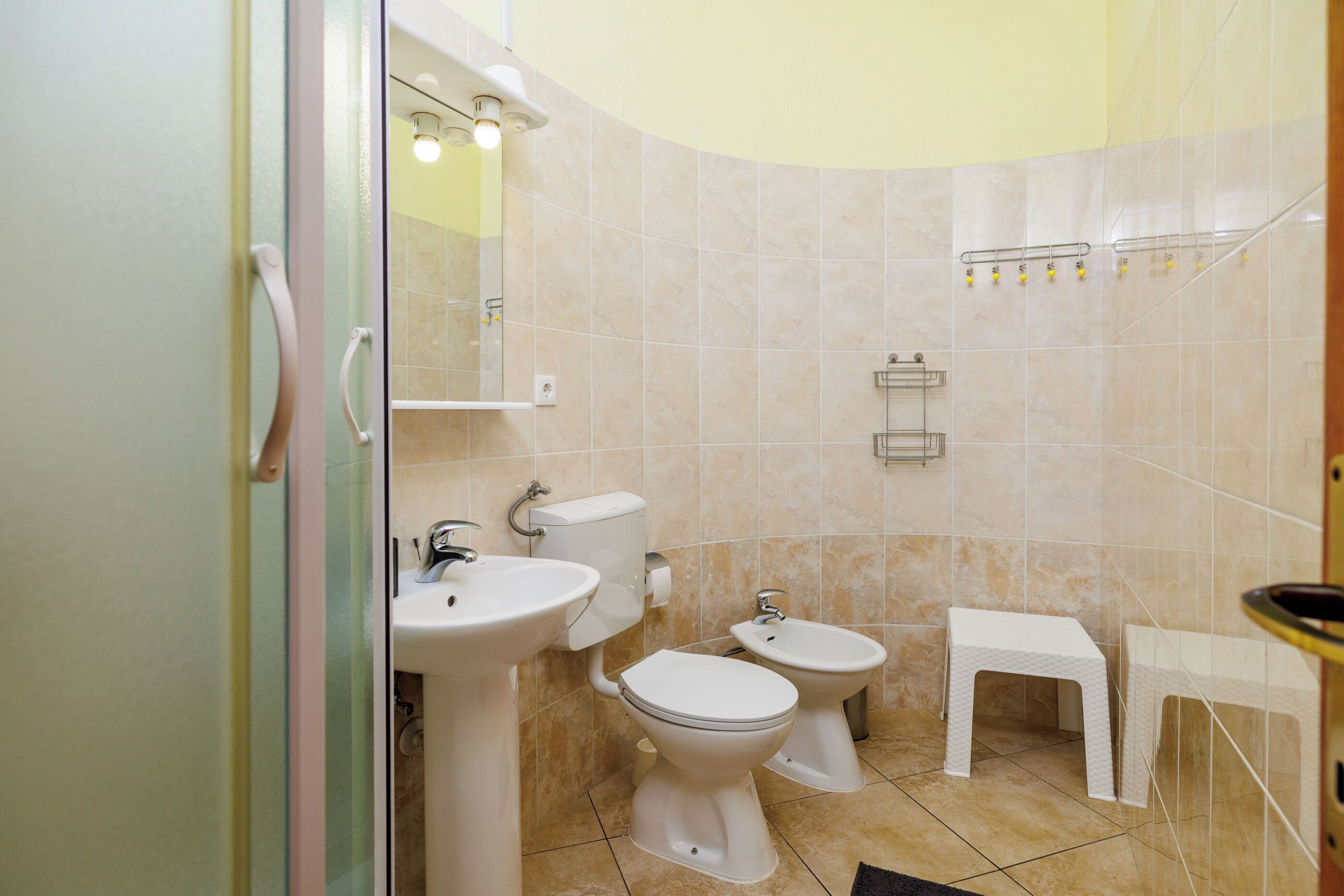 A bathroom with a toilet, bidet, sink, and a small stool.