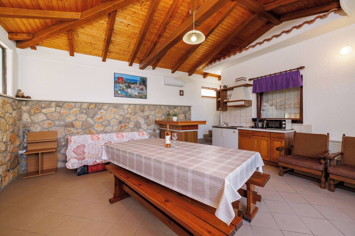 A cozy room with a wooden ceiling, a stone wall, a bed with a checkered bedspread, a wooden bench, and a dining table.