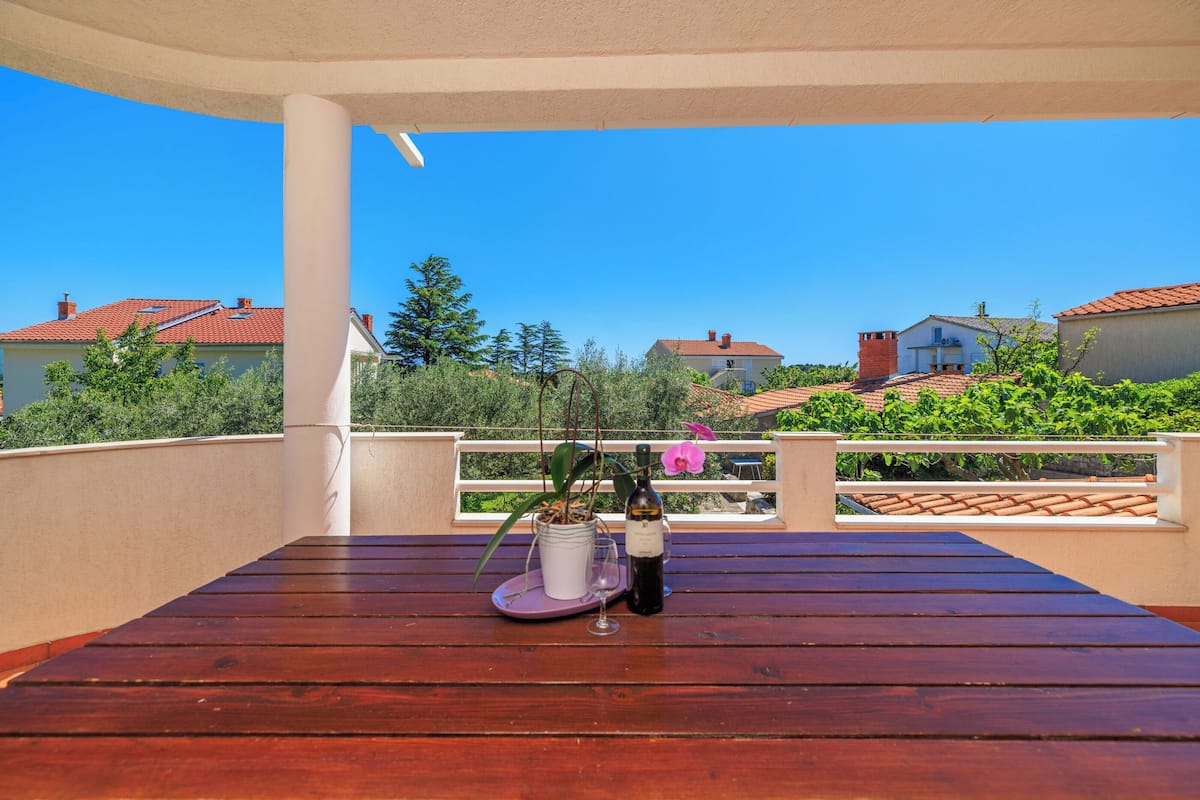 A wooden table with a wine bottle and glass on a balcony with a view of houses and greenery.