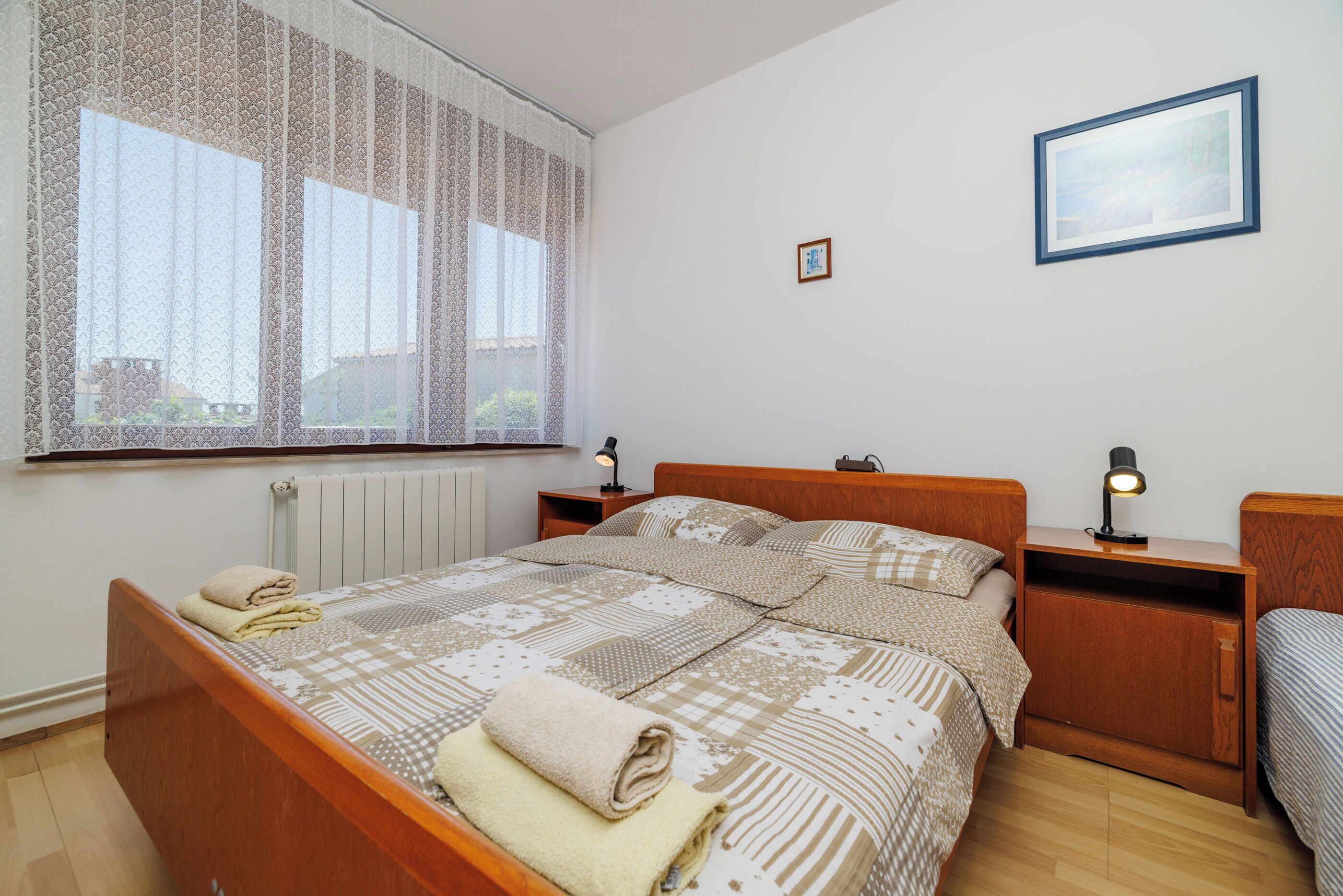 A bedroom with a large window, a bed with a patterned bedspread, two bedside tables with lamps, and a framed picture on the wall.