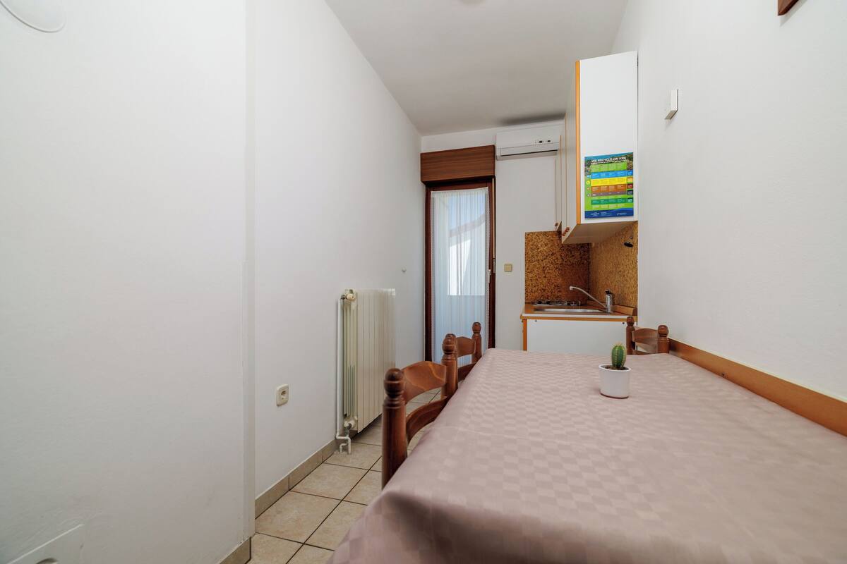 A small, compact room with a bed, a chair, a small table, a radiator, a window with curtains, a kitchen area with a sink and a stove, and a wall-mounted air conditioner.
