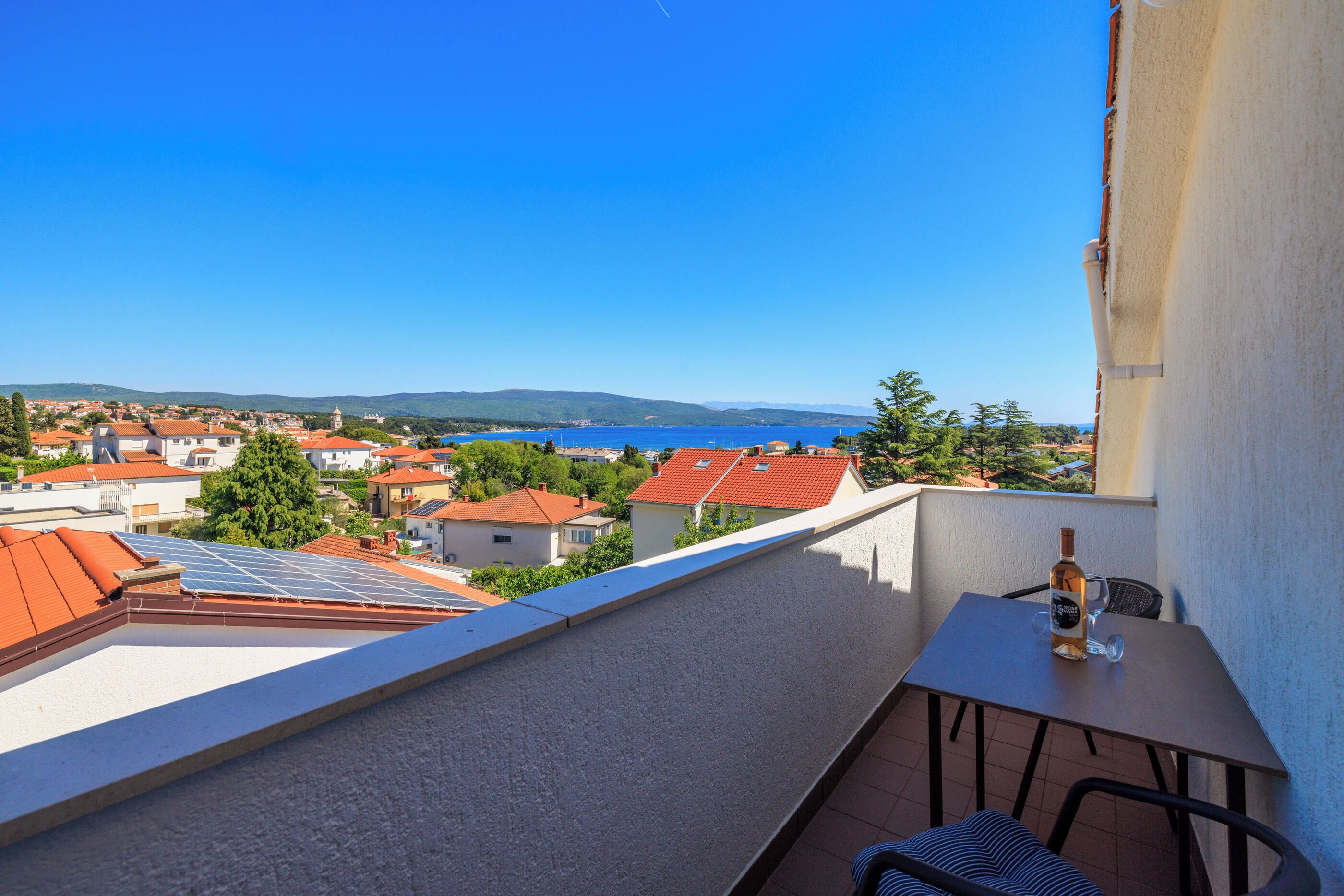 Un balcon avec une table et des chaises, donnant sur un quartier résidentiel avec des maisons aux toits rouges et un ciel bleu dégagé.