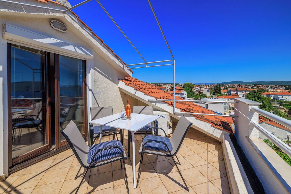 A balcony with a table and chairs, a bottle, and a view of a town with red-roofed buildings.