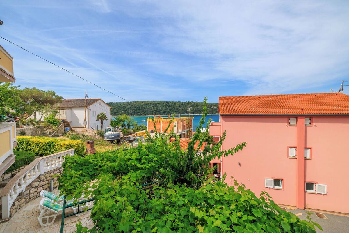 A coastal residential area with a pink building, a white building, and a yellow building. There is a staircase leading to a balcony with a chair.