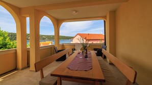 A balcony with a wooden table, benches, and a view of a building and landscape. - Apartments Ljubica (Kampor)