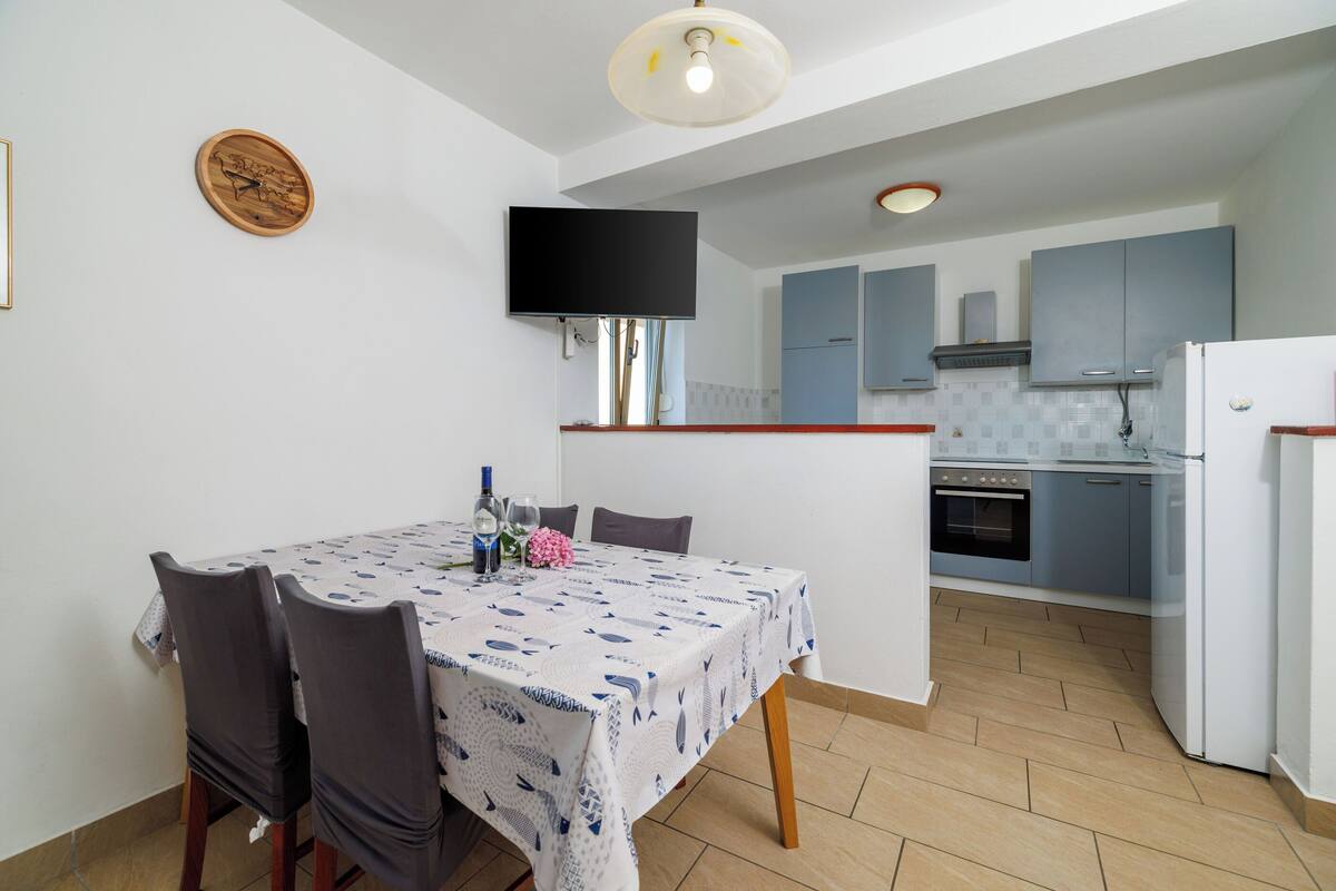A dining area with a table set for four, a television mounted on the wall, and a kitchen in the background.