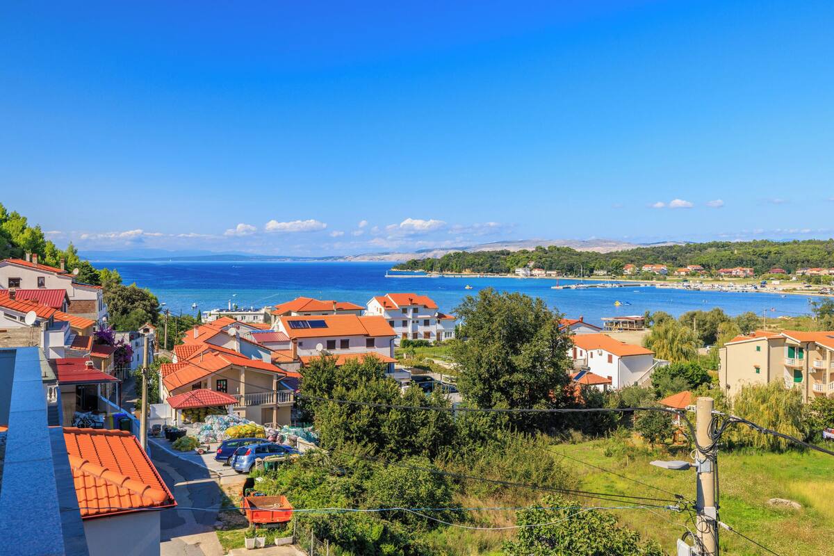 A coastal town with red-roofed buildings, a clear blue sea, and a hilly landscape in the distance.
