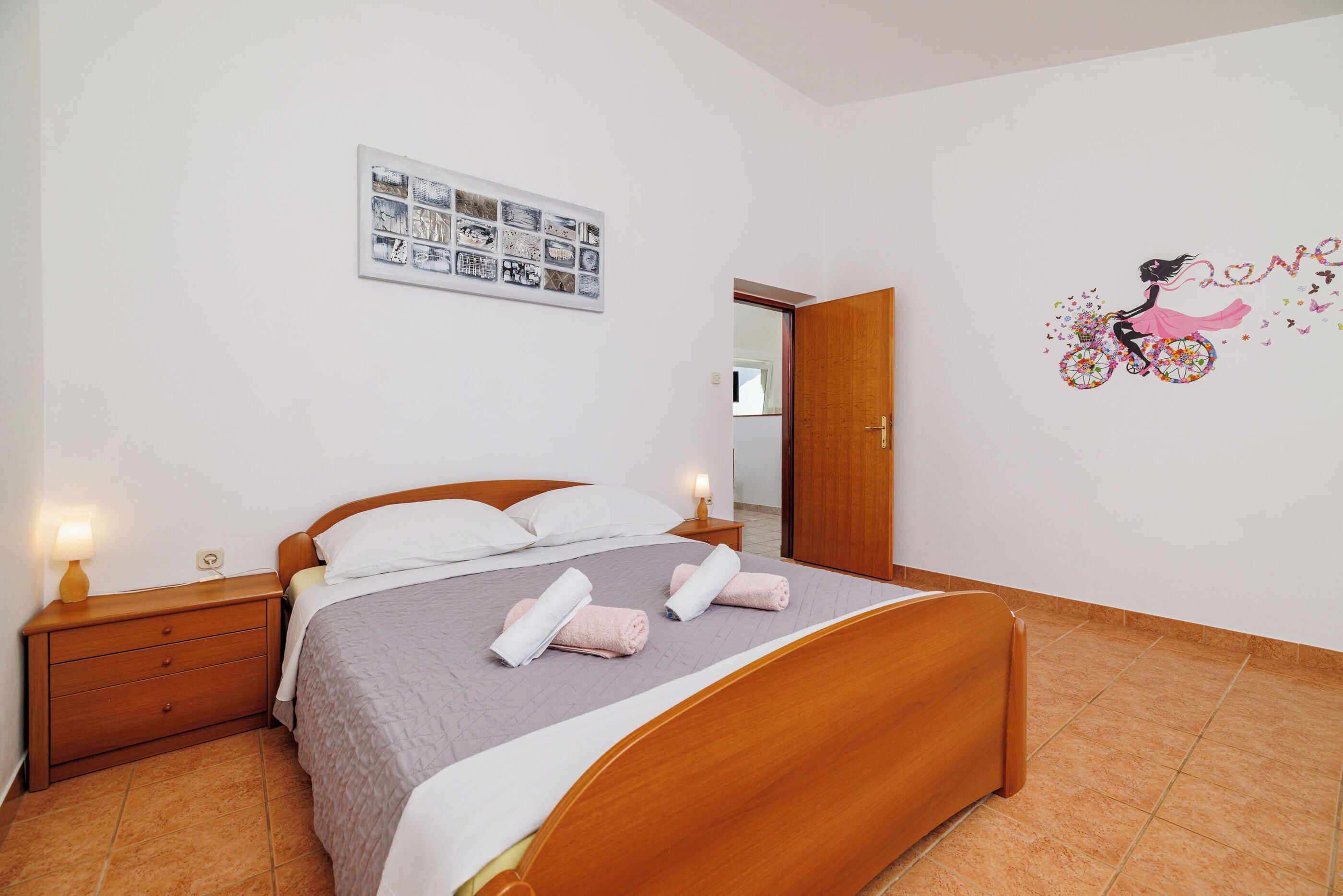 A neatly made bed with a headboard, a bedside table with a lamp, and a wall decoration of a girl on a bicycle.