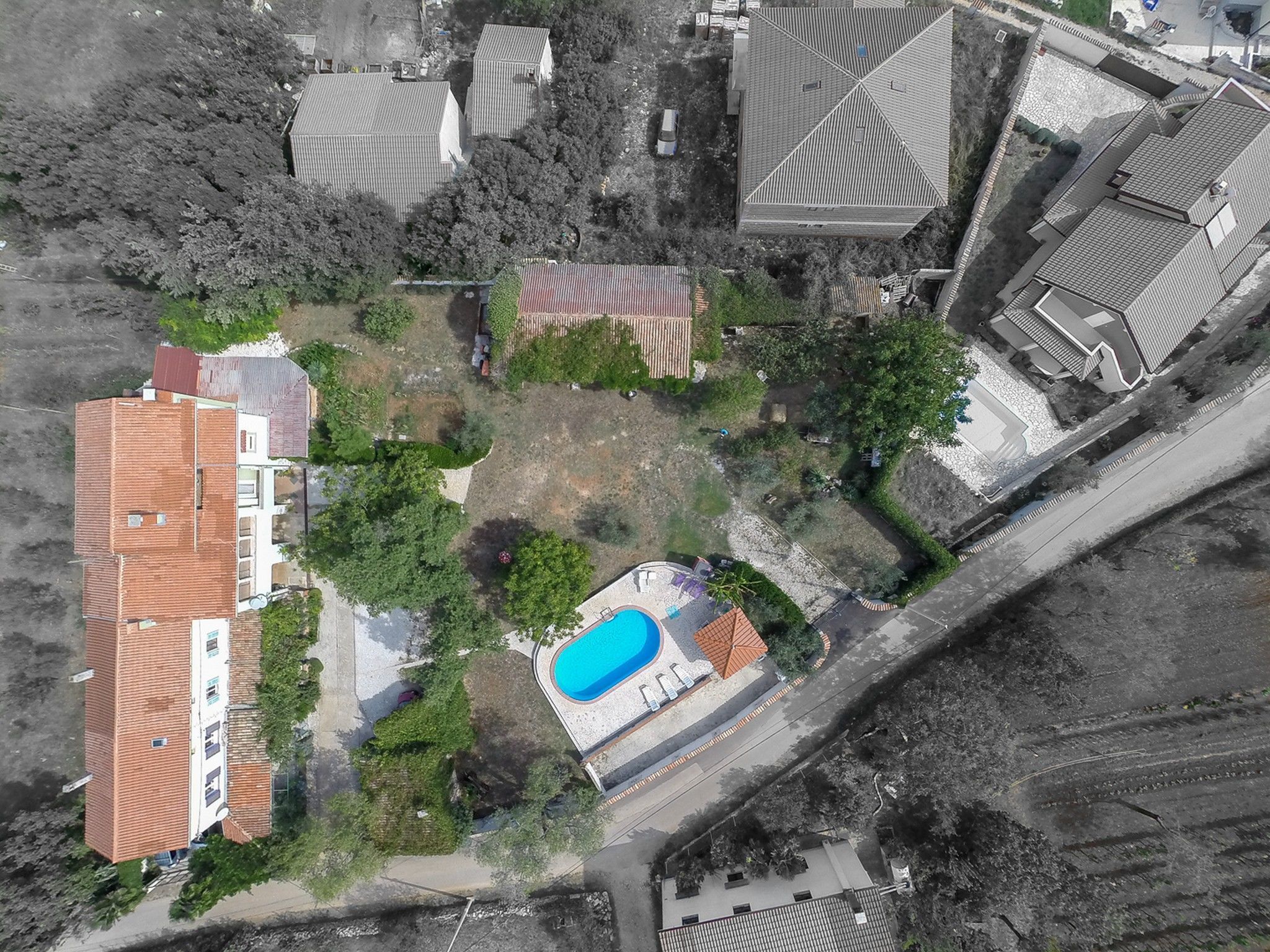 An aerial view of a residential area with a large building featuring a red-tiled roof, a swimming pool, and surrounding greenery.