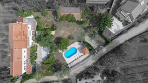 Vista aérea de um bairro residencial com um edifício grande, telhado de ladrilho vermelho, piscina e vegetação ao redor.