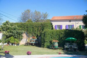 A house with a red-tiled roof, purple shutters, and a garden with a swimming pool.