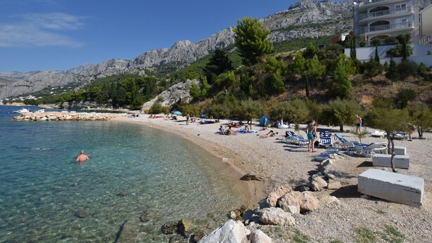 A beach with clear water, rocky shore, and a building with balconies overlooking the sea. - Apartment Anica (Omis)