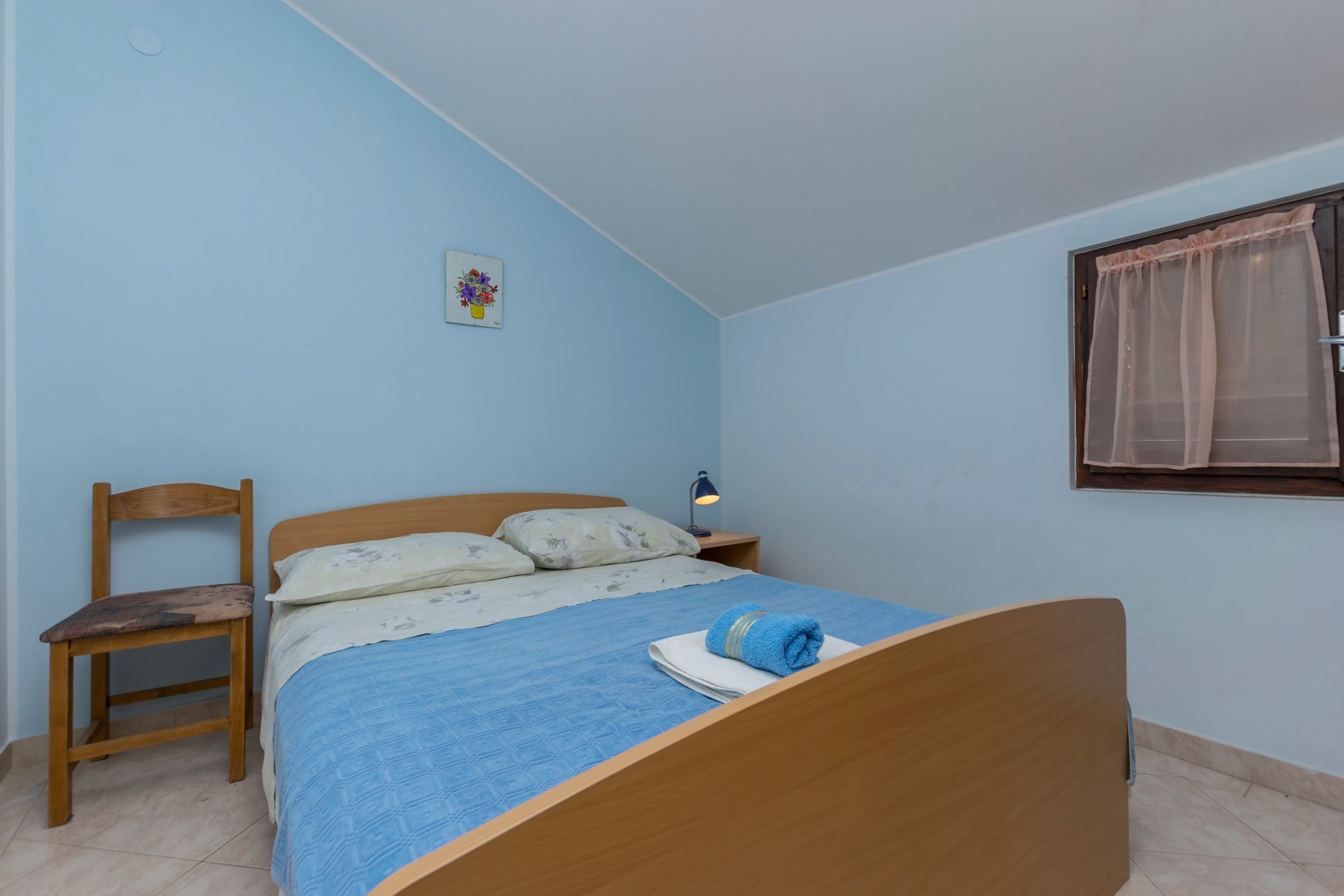 A bedroom with a wooden bed, a chair, a nightstand with a lamp, and a window with a sheer curtain.