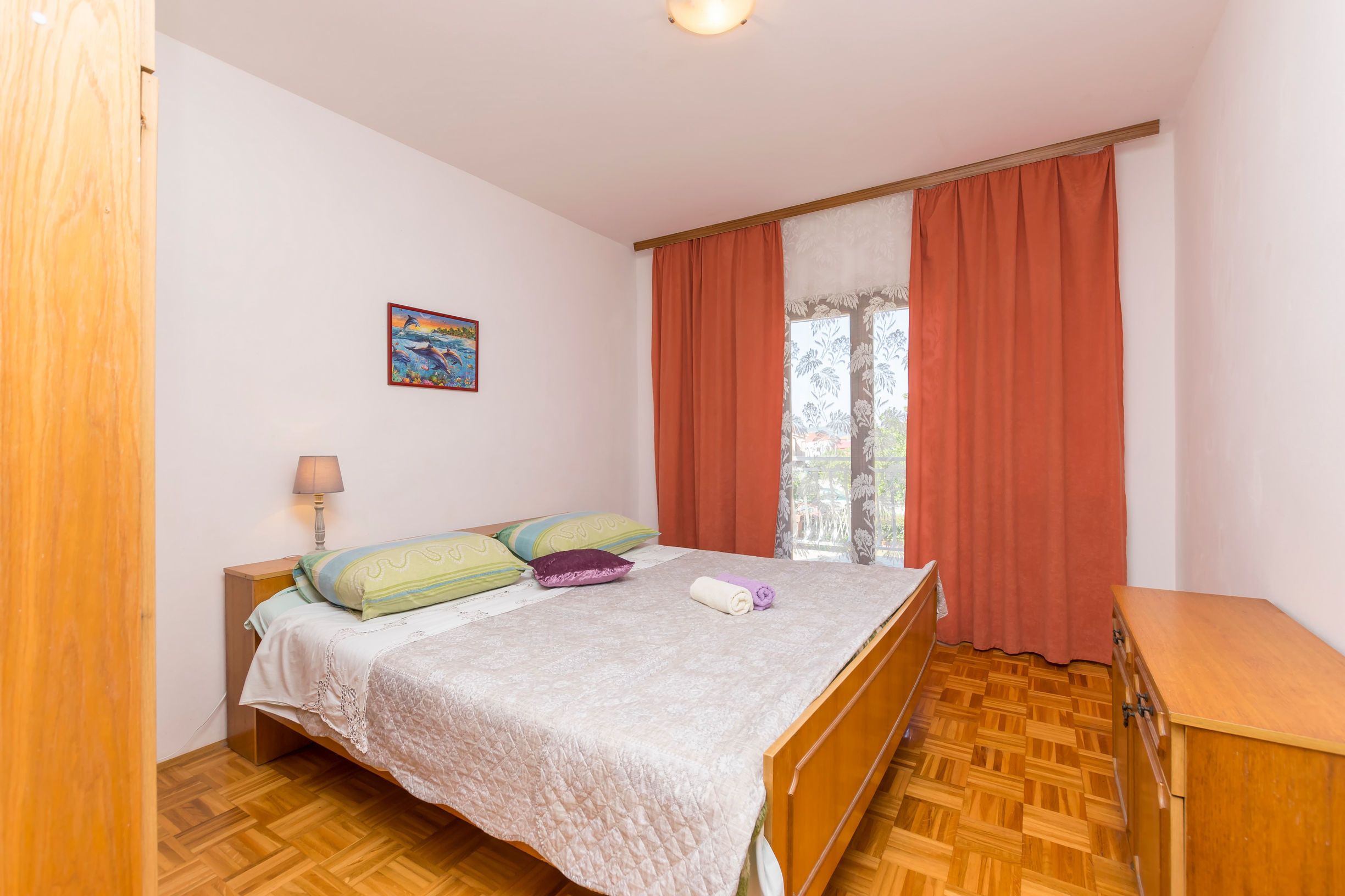 Une chambre à coucher comprenant un lit, une table de chevet, une lampe, une fenêtre avec des rideaux et une commode en bois.