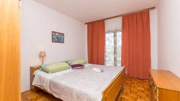Une chambre à coucher comprenant un lit, une table de chevet, une lampe, une fenêtre avec des rideaux et une commode en bois.
