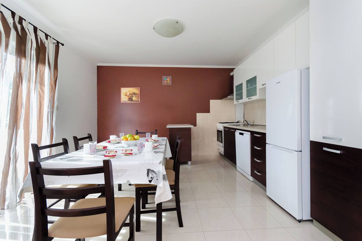 Un coin repas avec une table dressée pour quatre personnes, une cuisine équipée d’appareils électroménagers blancs et un escalier menant à l’étage supérieur.