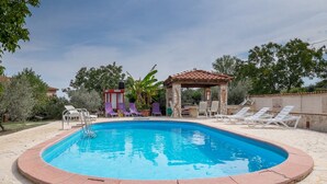Ein Schwimmbecken mit gefliestem Sonnendeck, umgeben von Liegestühlen und einer überdachten Sitzgruppe.