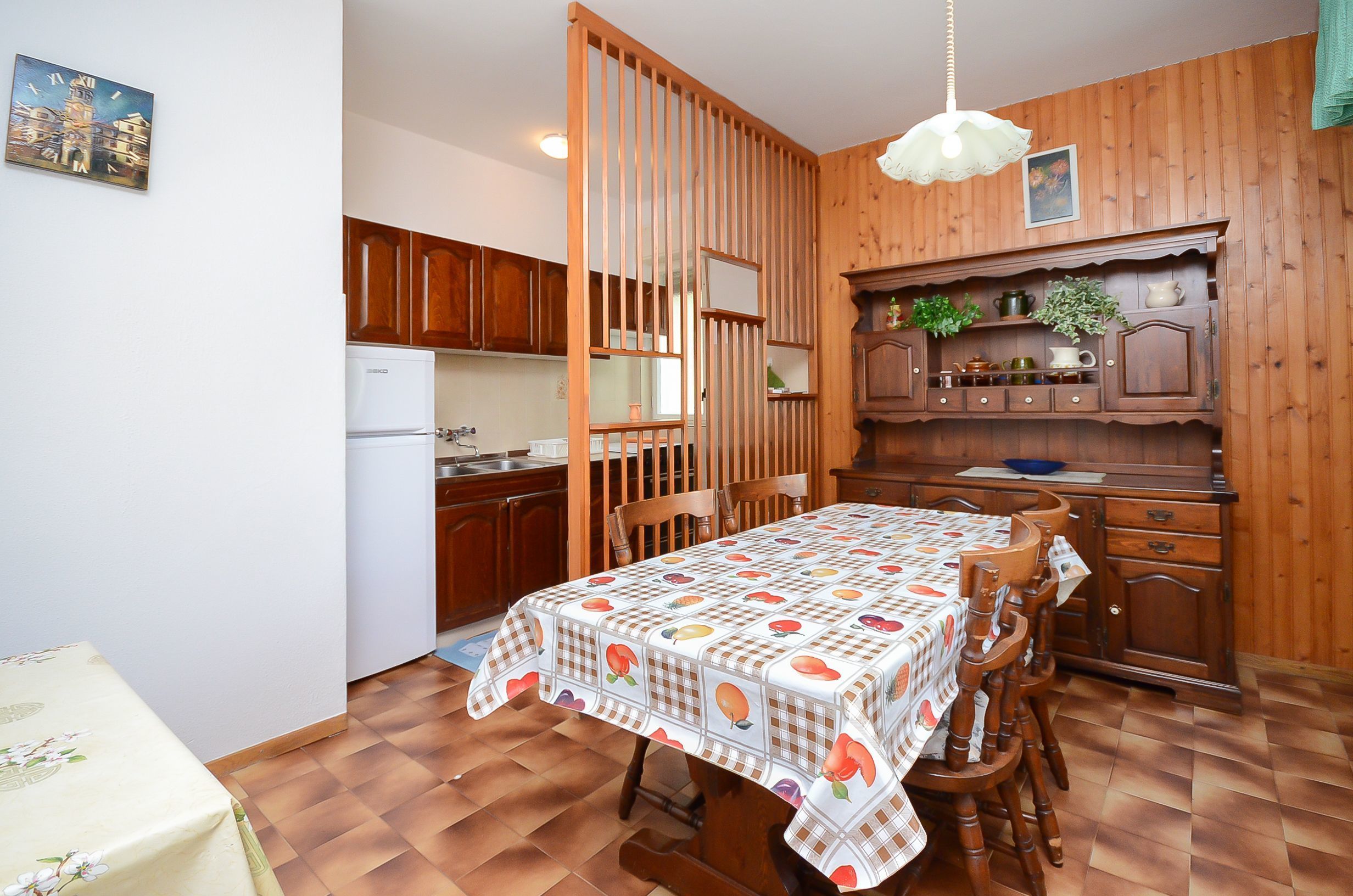 Eine kleine Küche mit Holzausstattung, einem Kühlschrank und einem Esstisch mit Obstmuster-Tischdecke.