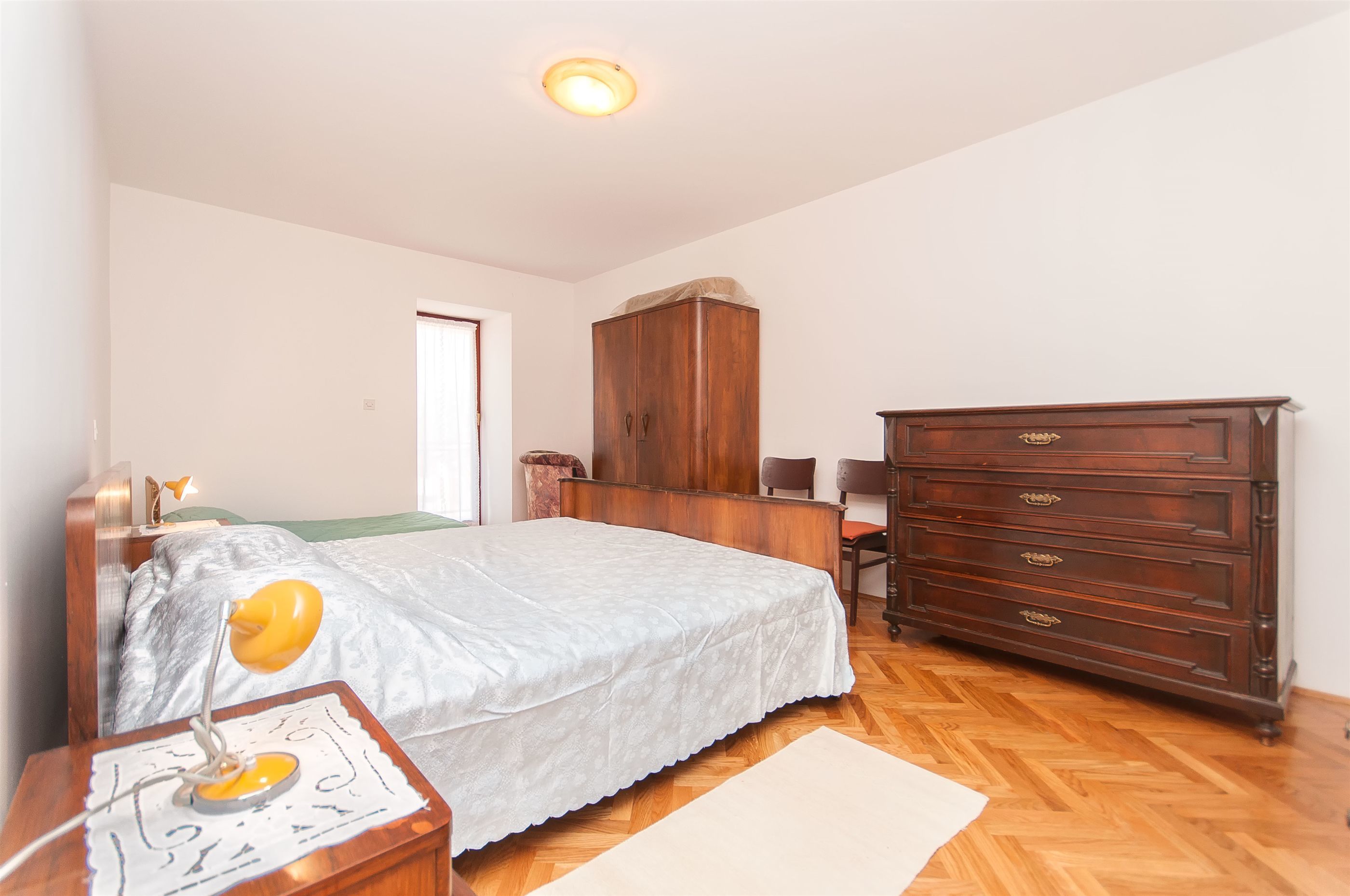 Een slaapkamer met een bed, een houten dressoir, nachtkastje en een lamp.