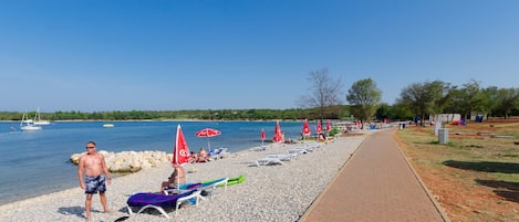 A pebbled beach with lounge chairs and umbrellas, a clear sky, and a calm body of water in the background.