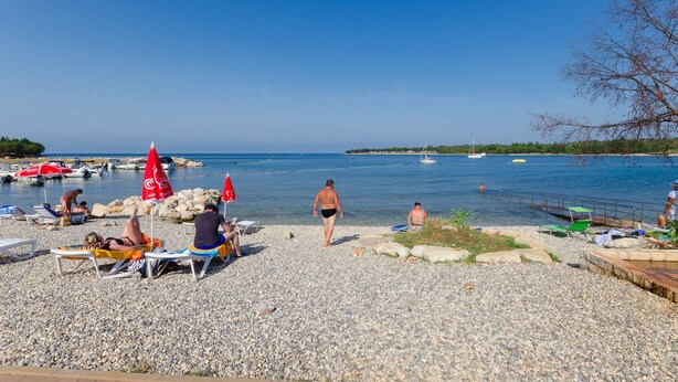 A pebbled beach with people sunbathing and swimming, boats in the water, and a clear sky. - Apartment Marija (Porec)